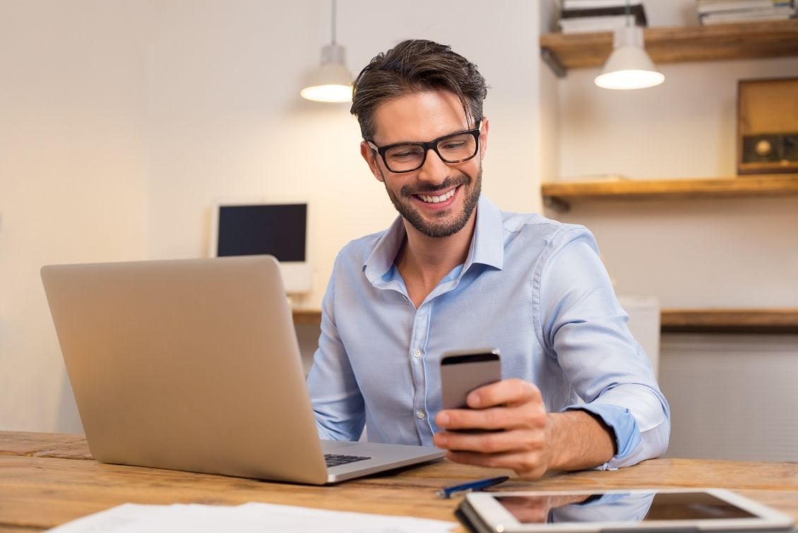 Man holds phone and smile as he sits in front of an opened laptop