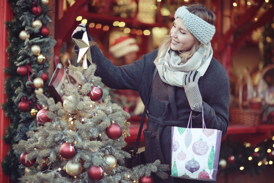 Woman in winter clothes touches the star on top of a Christmas tree as she holds a shopping bag