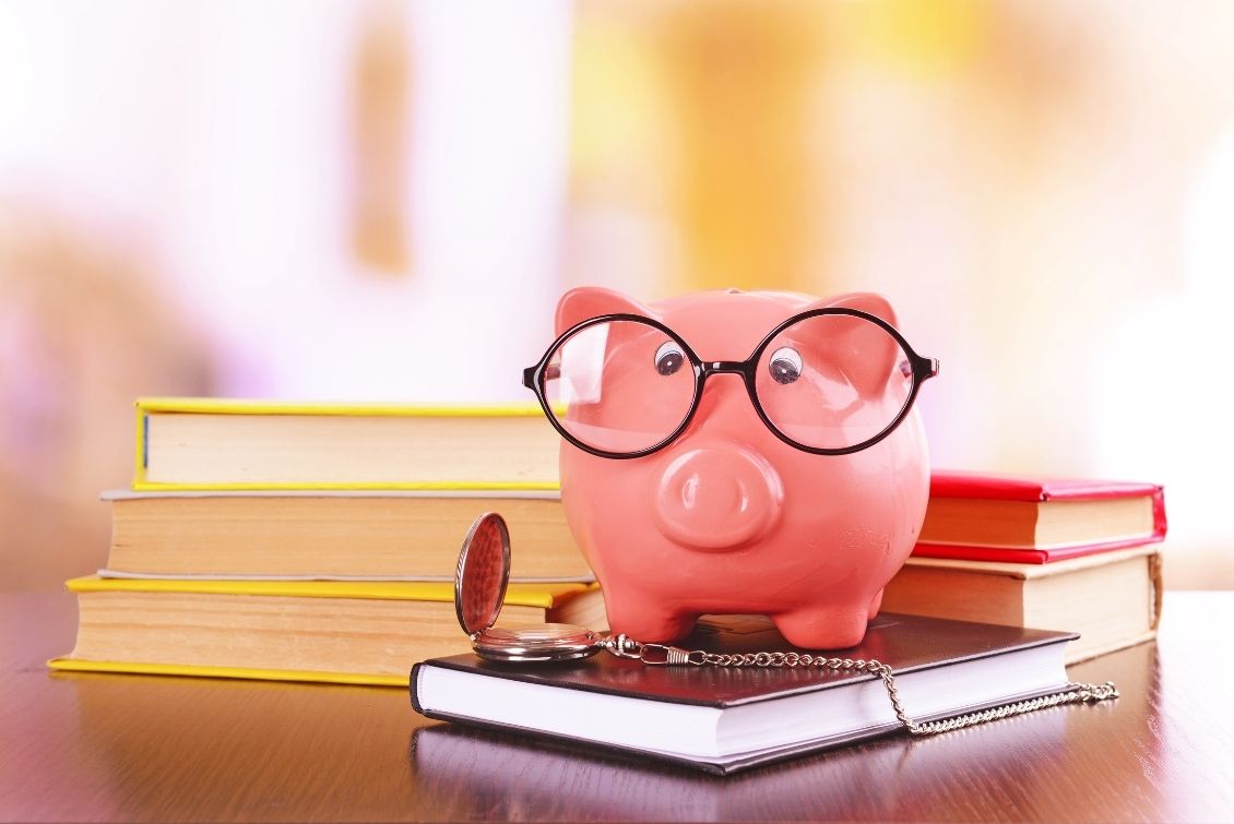 A piggy bank with glasses sits on top of a book with an open pocket watch next to it with stacked books in the background