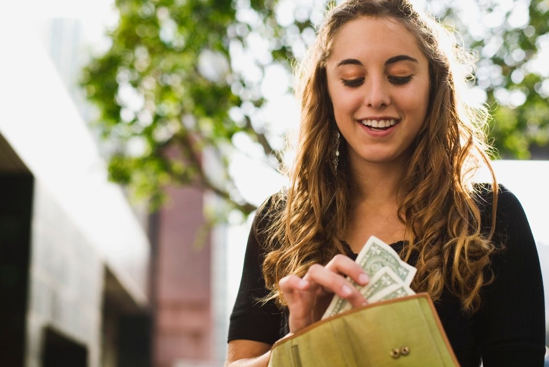 A smiling young woman is putting three dollar bills into a wallet