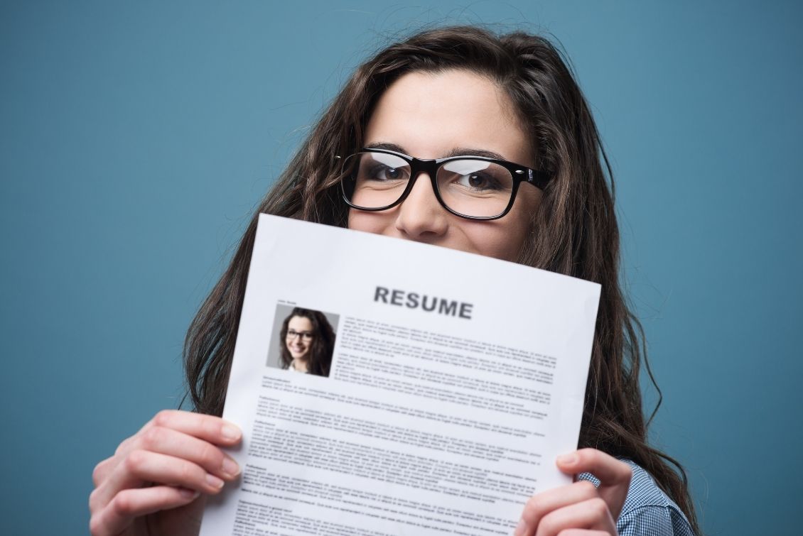 Young woman with glasses holds her resume in front of her face in front of a blue background