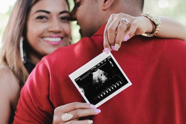A woman holds up a sonogram while she smiles and hugs a man in a red shirt