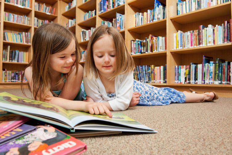 Two young girls at the library reading a book on the ground