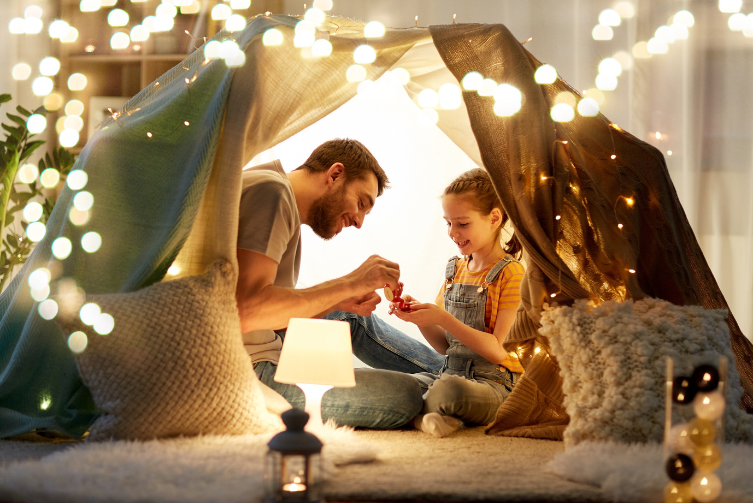 A father and a daughter under a fort made of blankets. He pours her a cup of tea.