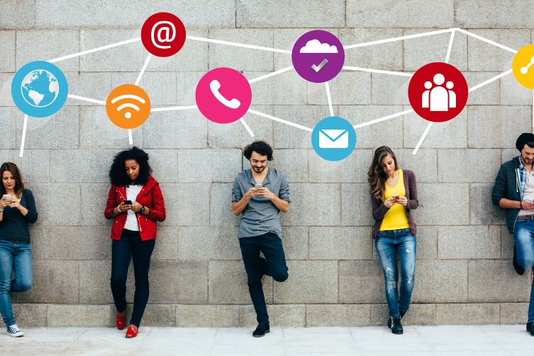 A group of adults lean against a wall using their phones with social media icons above them.