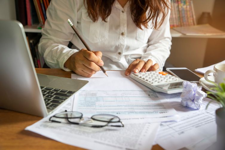 Woman is surrounded by financial paperwork, a calculator, a pair of glasses, a phone and laptop.