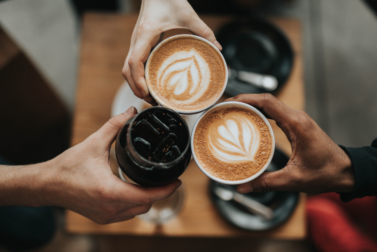 Three hands holding lattes (hot and cold) come together in the center of an overhead shot.