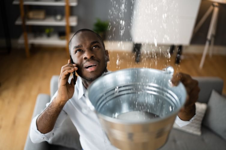 A man holds a bucket to catch water as he holds a phone to his ear with his other hand..