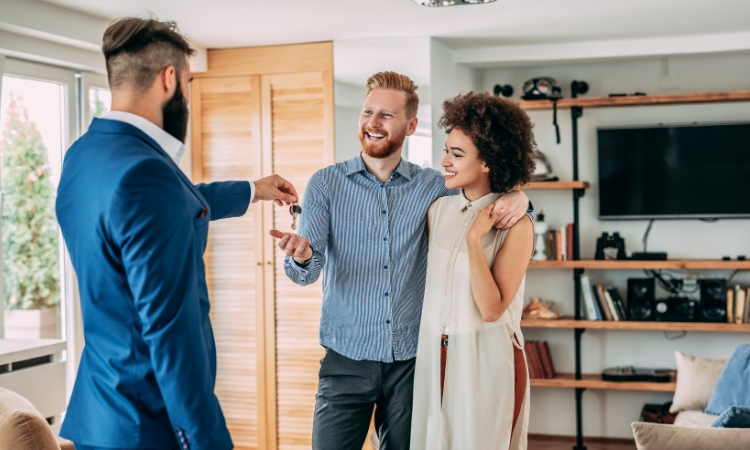 A young man and woman smile as a realtor hands them a key to their new home.